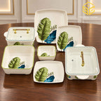 Glamour botanical plates, bowls and covered serving dishes with gold-tone handles are arranged on a wooden table.