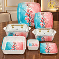 Glamour turquoise and pink blossom plates, bowls and covered serving dishes with gold-tone handles are arranged on a wooden table.
