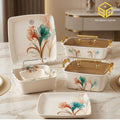 Modern floral plates and bowls accompany two amber-lidded serving dishes with gold-tone handles on a cream marble table.