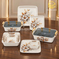 Modern floral plates, nested bowls and two covered serving dishes are arranged on a wooden table.